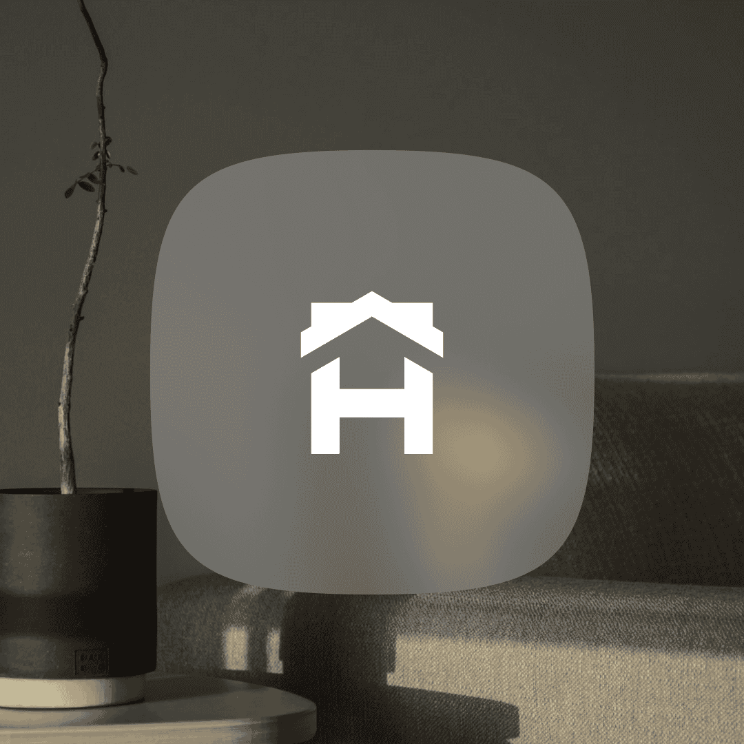 A white house icon with a roof and an "H" shape is centered on a gray rounded square, placed over a blurred background of a couch, wall, and potted plant.